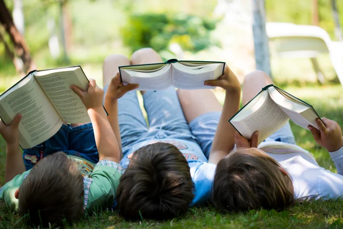 Photo of three children lying on their backs on a grass lawn & reading books.