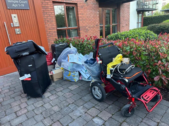 Image shows a red and black powerchair, three black suitcases and a pile of plastic bags and boxes left outside an apartment 