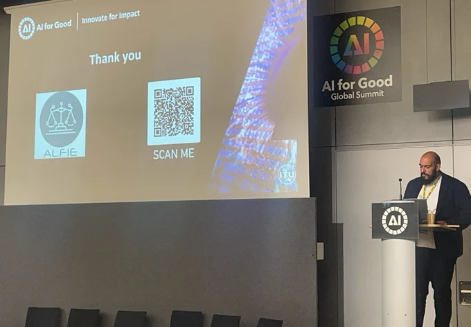Zenon Lamprou is presenting on stage, standing at a podium with a large screen behind displaying the ALFIE project logo and a QR code.