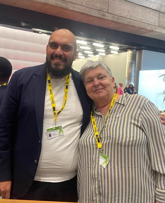 Pilar Orero and Zenon Lamprou smiling together at the AI for Good Global Summit, both wearing event badges and yellow lanyards.