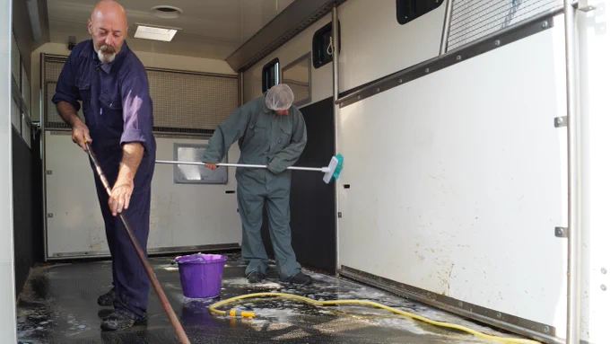 A photo of two people in overalls scrubbing and cleaning a horse box.