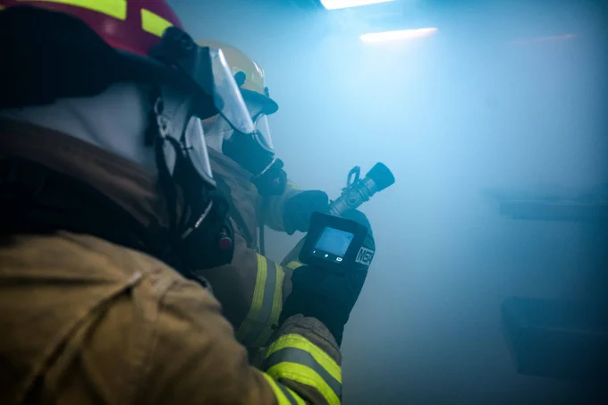 Two firefighters in full gear, including helmets and face masks, operate inside a smoke-filled room. One holds a thermal imaging camera, while the other grips a fire hose nozzle. The scene is bathed in blue overhead lighting, enhancing the dramatic atmosphere.