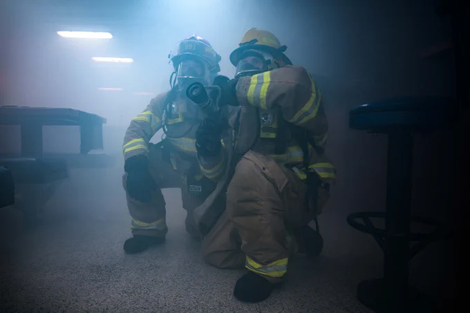 Two firefighters in full gear, including helmets and breathing apparatuses, kneel on the floor of a smoke-filled room. One holds a hose nozzle, ready to spray water. Tables and chairs are partially visible through the thick smoke, and dim lighting enhances the hazy atmosphere.