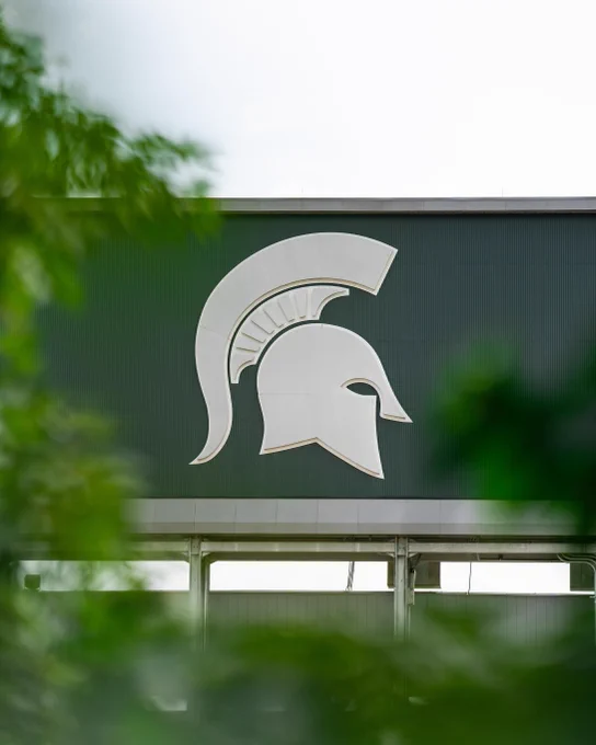 Spartan helmet emblem on a dark green building, with blurred green leaves in the foreground.