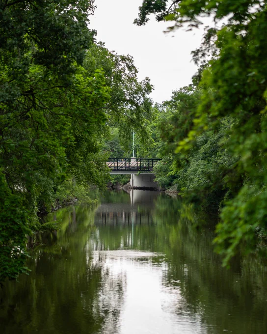 A river with green trees on both sides and a footbridge in the distance.