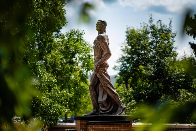Statue of a Spartan warrior draped in flowing garments, set against a backdrop of green trees and white sky.