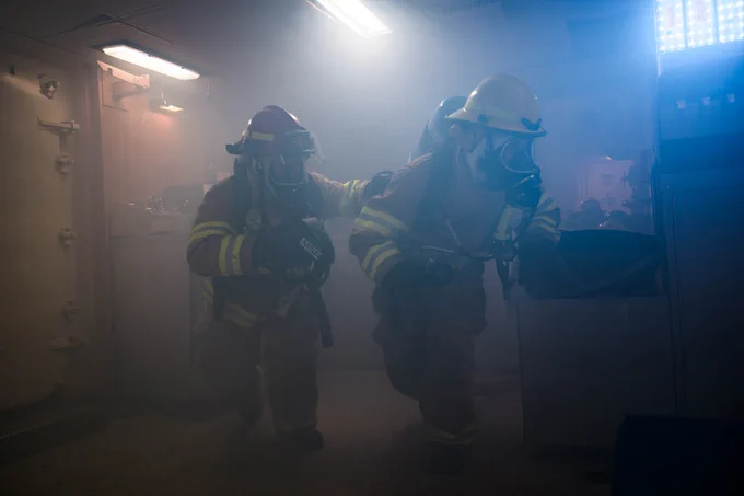 Two firefighters in full gear, including helmets and breathing apparatus, move through a smoke-filled room. The firefighter in front wears a yellow helmet and leads the way, while the one behind, in a purple helmet, holds onto their shoulder. Dim lighting and scattered equipment are visible through the haze.