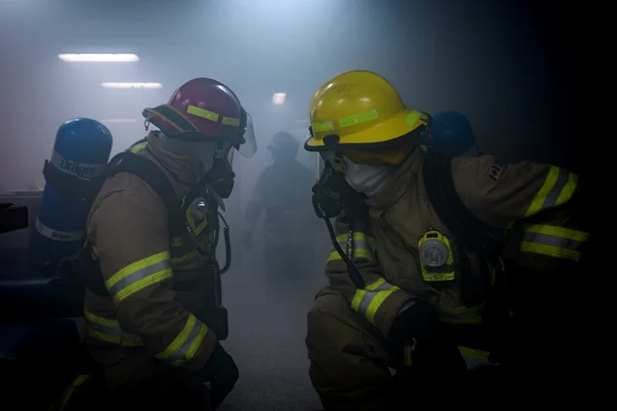 Two firefighters in full gear, including helmets, masks, and oxygen tanks, operate in a smoke-filled indoor environment. The firefighter on the left wears a red helmet, and the one on the right wears a yellow helmet. Both have reflective stripes on their suits, with a third firefighter faintly visible in the background through the smoke.