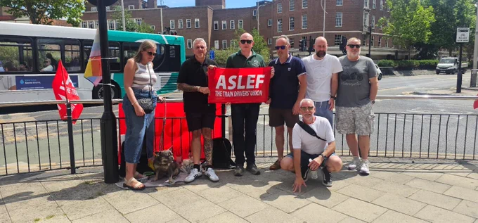 Group of people standing together with ASLEF flags on an official picket line. 