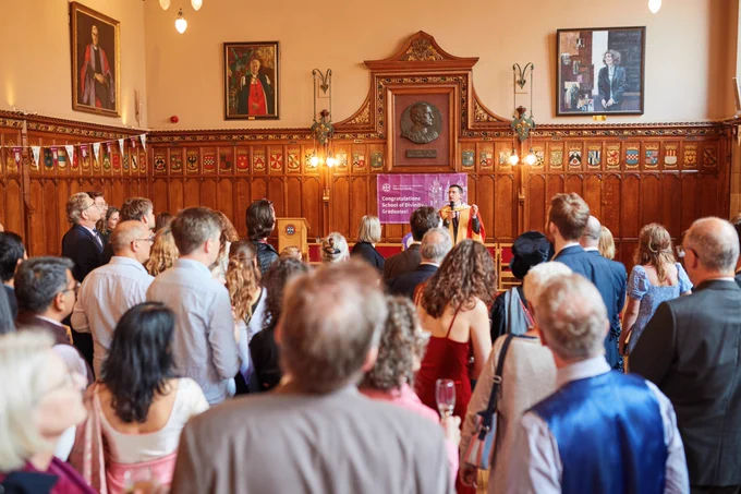 Colour photo of Professor Jeremy Carrette stood on the Rainy Hall stage speaking towards attendees of the School of Divinity graduation celebration who are facing him. The photo is taken from behind the guest's head.