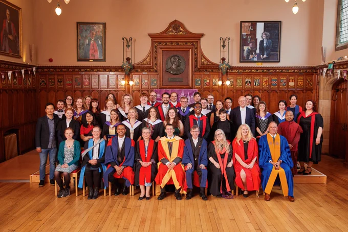 Colour photo of School of Divinity graduating students and staff stood and seated in the Rainy Hall smiling towards the camera