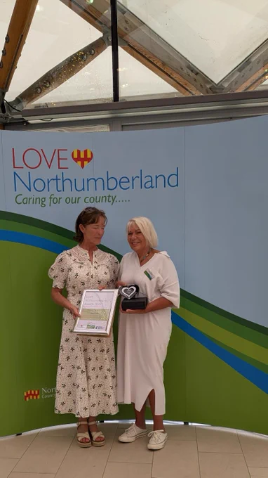Two women stand in front of a backdrop with "LOVE Northumberland" and "Caring for our county" text. One woman in a floral dress holds a framed certificate; the other, in a white dress, holds a black box with a heart-shaped award. They are inside a building with wooden and glass roofing.