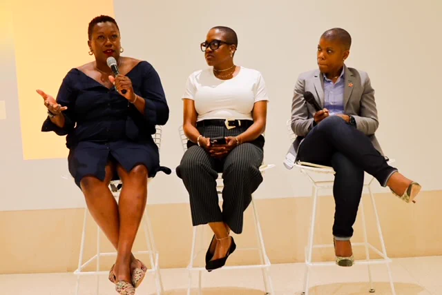 Three panelists sit on tall chairs in front of a white wall. The person on the left is speaking into a microphone. All are dressed professionally and appear engaged in the discussion.
