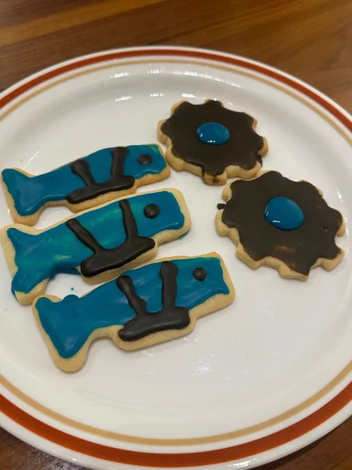 A photo of sugar cookies on a plate. Three of the cookies look like whales with brown harnesses, while the other 2 are brown gears with a blue circle in the middle.