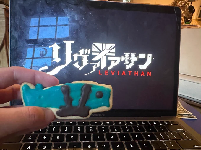 A photo of a sugar cookie shaped like the Leviathan, a whale airship with a harness and gondola, in front of a laptop playing the new Leviathan anime.
