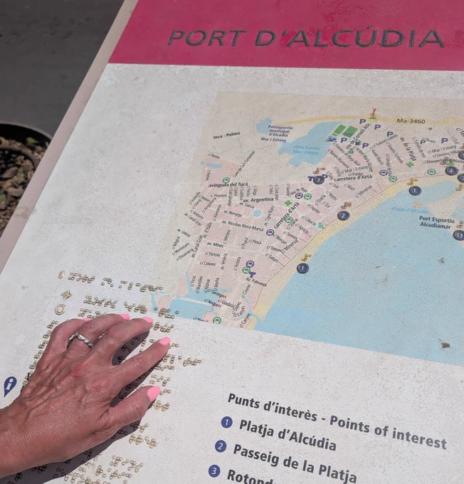 A hand with pink painted nails touches a tactile map of Port d’Alcúdia, Mallorca. The map includes braille and raised elements to help blind and visually impaired people navigate. Points of interest are listed in both text and braille.