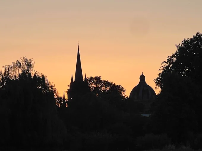 Silhouette of Oxford’s skyline at sunset, featuring the spire of the University Church of St Mary the Virgin and the dome of the Radcliffe Camera, framed by trees against a soft orange sky.