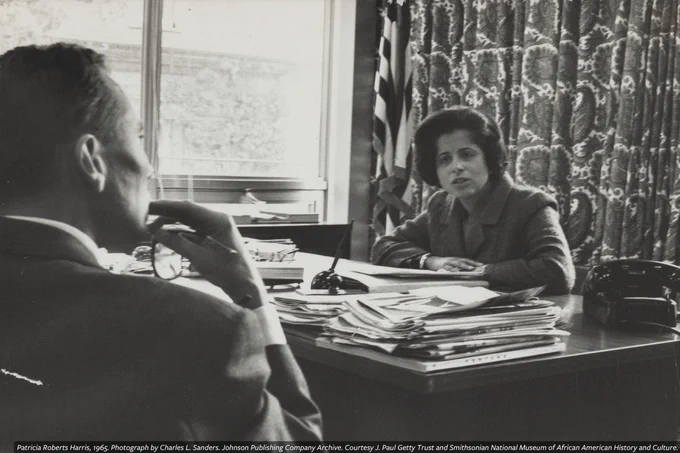 A black-and-white photograph of Patricia Roberts Harris as she sits at a desk piled with documents, listening attentively to Lyndon B. Johnson, who is facing away from the camera, in an office setting.