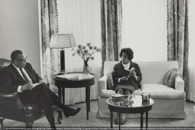 A black-and-white photograph of Patricia Roberts Harris and a man sitting in an elegant room, engaged in a discussion. Harris is reading a document while he listens attentively.