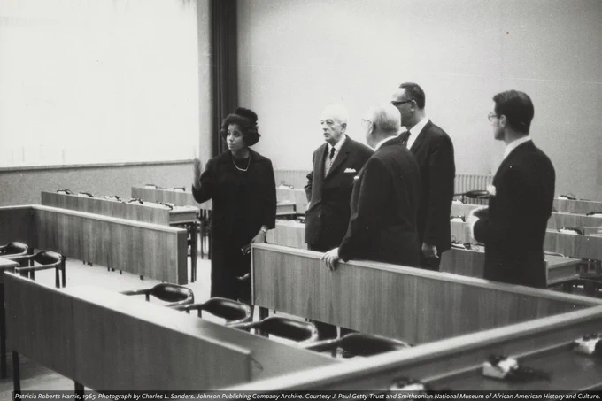 A black-and-white photograph of Patricia Roberts Harris speaking to a group including three men in a lecture hall, in a photograph by Charles L. Sanders.
