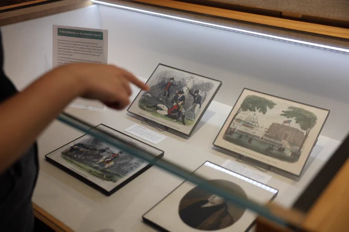 A hand is pointing to an image in a glass case showing men at battle.