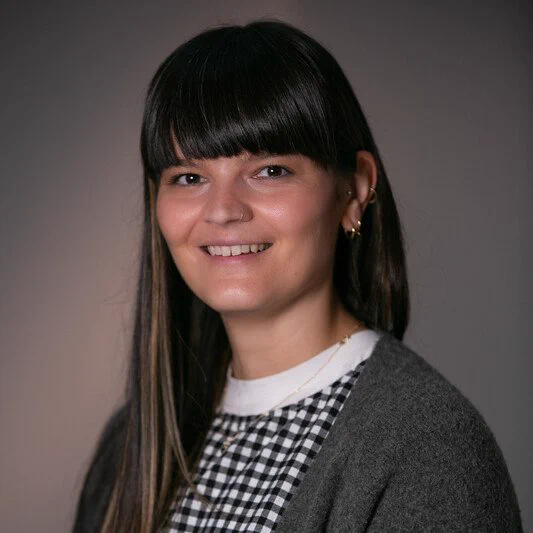 A portrait photo of Dr Lauren Lucas. Lauren has long dark hair and she is wearing a black and white top with a grey cardigan. 