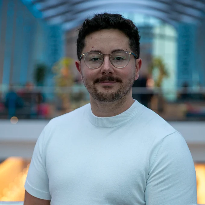 A portrait photo of Dr Sam Webb. He has dark hair, glasses and he is wearing a white t-shirt. 