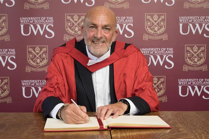 A person in a red academic robe is seated at a table, smiling while signing a book. The backdrop displays the University of the West of Scotland logo repeatedly.