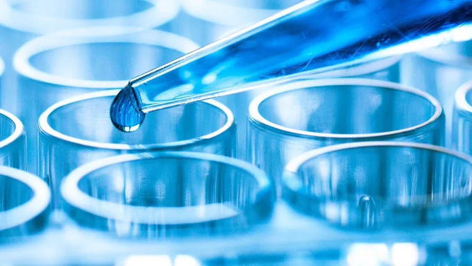 Close-up of a blue liquid drop being dispensed from a pipette into a row of empty laboratory test tubes, highlighting scientific research or medical testing.