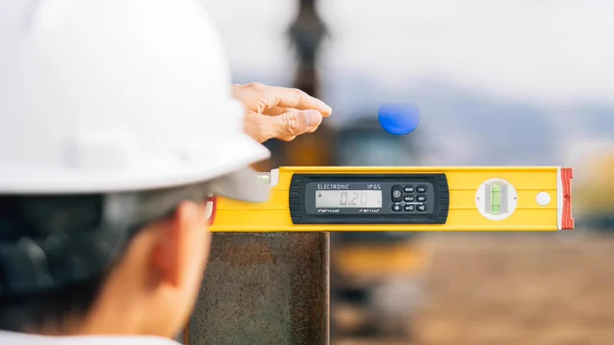 Person using a digital level tool to measure the angle of a metal beam on a construction site.