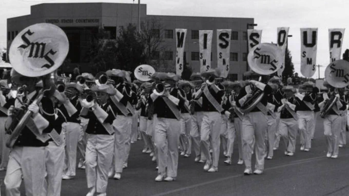 Marching band in uniform performing at an event, carrying banners that spell "MISSOURI" and symbols marked "SMS".