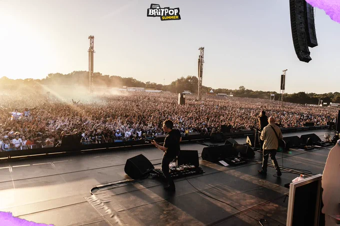 Photo from the back of the stage looking towards the audience, showing Oasis playing as the sun sets over a huge crowd in Manchester's Heaton Park