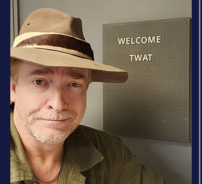 Rhys Darby dressed as Bill Napier in a bush hat and green shirt and silver chain stands smirking in front of a sign that says Welcome Twat