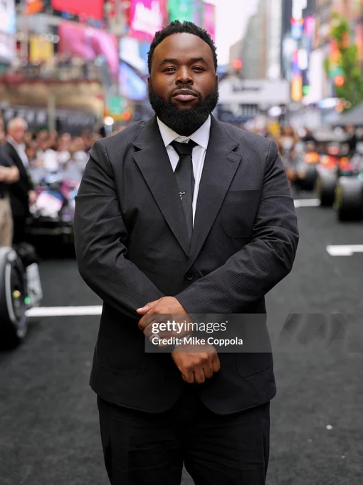 Samson Kayo stands in a street at the F1: The Movie NYC premiere in a black shirt with white Button down and black tie. Photo Ref says "GettyImages: Credit: Mike Coppola"