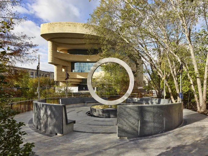 View of the National Museum of the American Indian, featuring a circular stone sculpture, the Native Veterans Memorial, in the foreground surrounded by lush greenery.