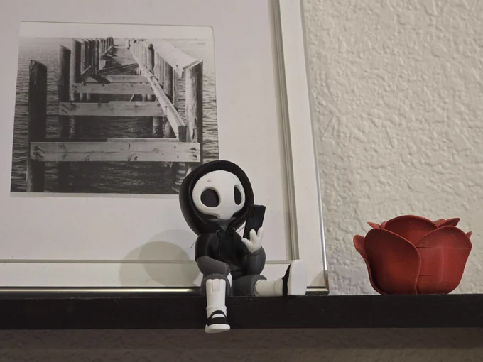 Project Hazy posed on a dark shelf between a framed photo of a pier and a red rose planter, sitting with feet dangling.