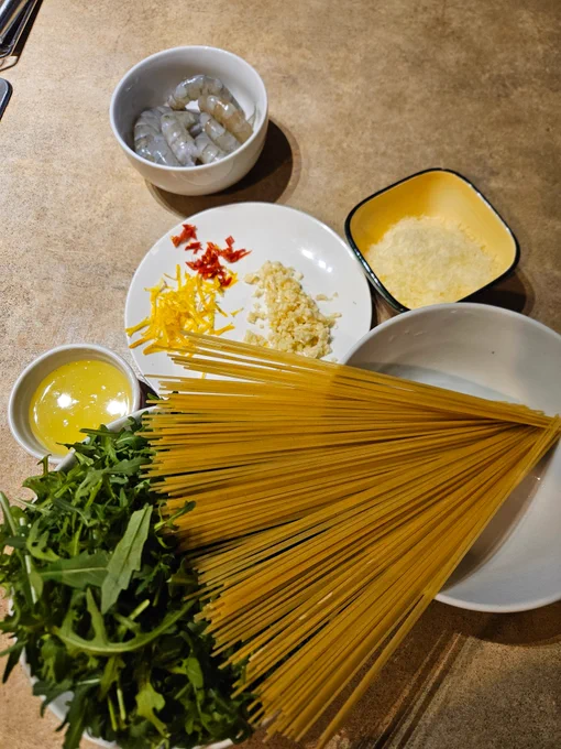 Ingredients clockwise from top of a small bowl of raw prawns, a ramekin with grated cheese, a bowl with dry spaghetti, a bowl of baby rocket leaves, a small bowl of lemon juice, a plate with lemon zest, minced garlic, sliced red chilli.