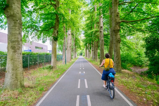 People in casual summer attire ride bicycles along a continuous regional cycling route in natural and built-up areas between Utrecht and Amersfoort.