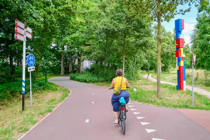 People in casual summer attire ride bicycles along a continuous regional cycling route in natural and built-up areas between Utrecht and Amersfoort.