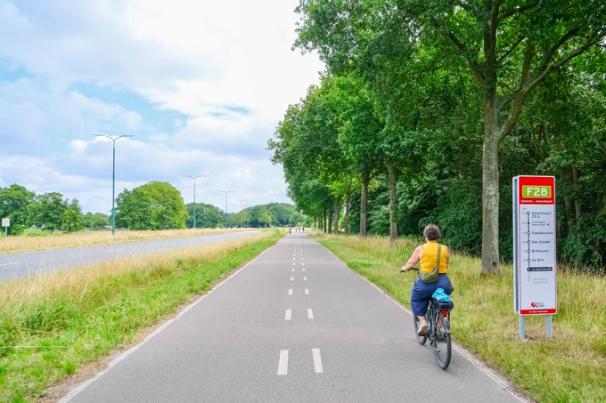 People in casual summer attire ride bicycles along a continuous regional cycling route in natural and built-up areas between Utrecht and Amersfoort.