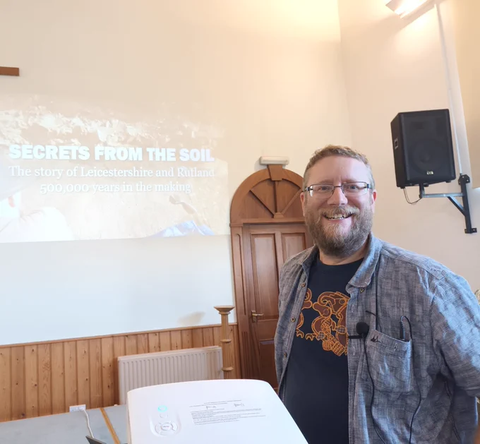 A smiling bearded man in a blue ahiet over a blue t shirt. Behind him is a white wall with words projectes on