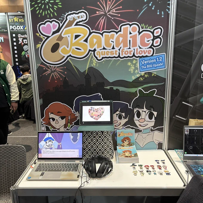 this is a photo of the PGDX booth for Bardic: Quest for Love by Breadwork Games. there are two screens on the table, some art books, and stickers.