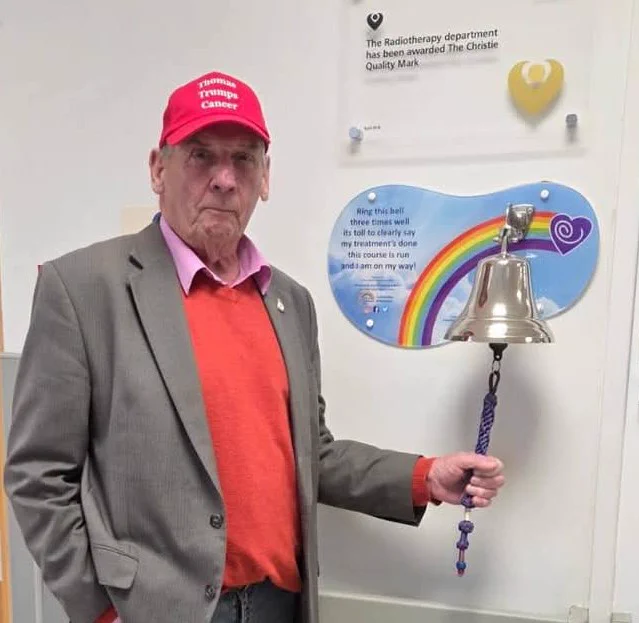Individual standing next to a commemorative bell and a plaque at The Christie Hospital, wearing a red cap and gray blazer.