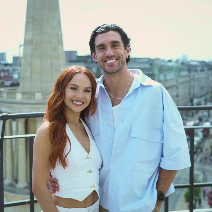Alexis Warr and Julian Caillon smile to the camera from a rooftop in Central London. A city skyline is visible in the background behind them. 
