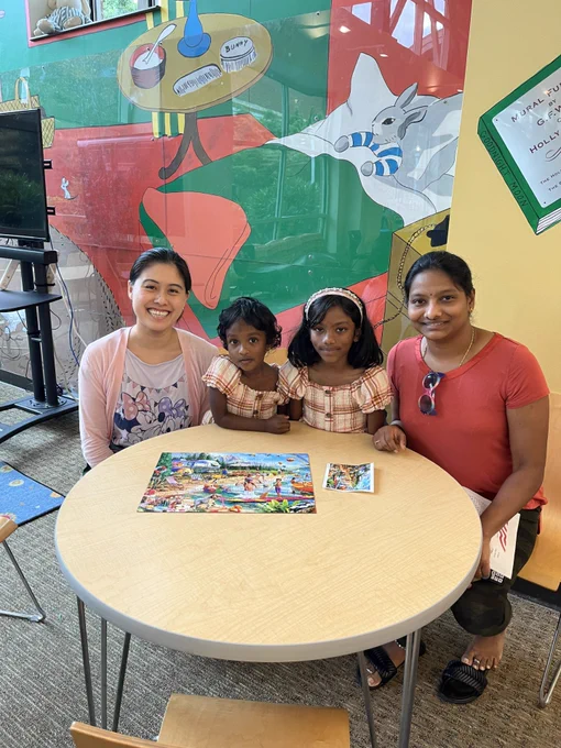 Two young girls in matching plaid dresses sit at a round table with two women, completing a colorful puzzle in a vibrant room with a mural of a rabbit and household items.