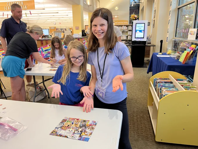 Two girls proudly display a completed colorful animal-themed puzzle on a table in a library setting.