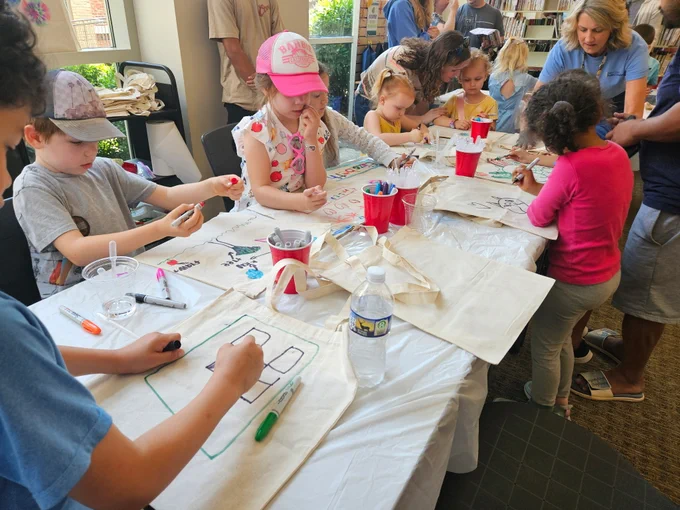 Children seated around a table drawing and coloring on canvas tote bags with markers during a group art activity indoors.