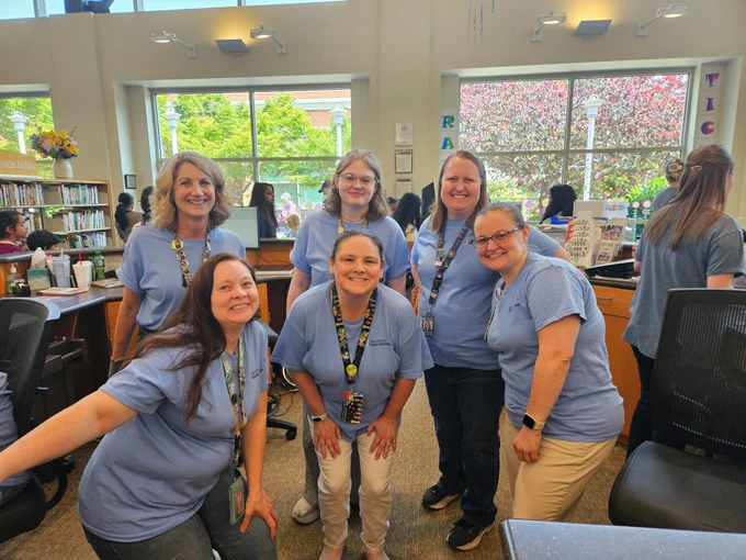 Six women wearing matching light blue shirts and lanyards pose together inside a bright library with large windows and bookshelves.