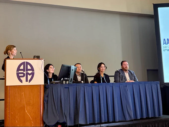 A woman standing behind an AAPM podium and giving a talk with a group of four moderators sitting behind a table on stage.