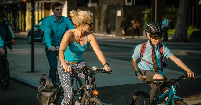A woman in a turquoise tank top and patterned pants rides a bicycle alongside a child in a school uniform and helmet on a city street.
